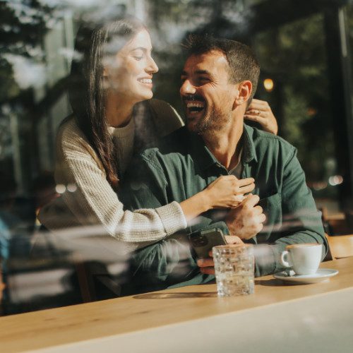 Young couple shares tender moments at a café, surrounded by greenery and natural light, as they connect over warm drinks and playful gesture